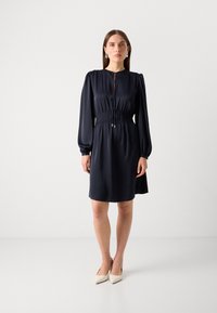 Navy blue dress made of smooth fabric, featuring long sleeves, a gathered waist, and a keyhole neckline. Paired with beige pointed shoes.