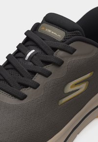 Σκούρο γκρι αθλητικό παπούτσι Skechers GOwalk με μαύρα κορδόνια, ύφασμα από δίχτυ, επενδυμένο γιακά και κίτρινο λογότυπο "S" στο πλάι.