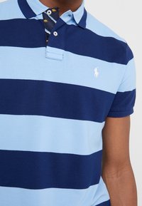Camisa polo a rayas azul y marino hecha de un tejido texturizado; presenta un cuello sólido en azul claro y una tapeta interior con un patrón y pequeños detalles.