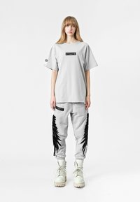 Camiseta gris de gran tamaño con etiqueta gráfica negra, combinada con pantalones joggers grises con detalles de flecos negros y botas blancas con cordones.