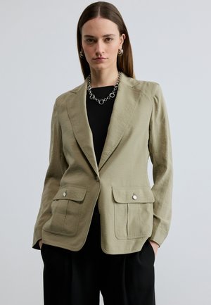 Femeie cu păr drept castaniu poartă un sacou culoarea măslin deschis cu buzunare aplicate, bluză neagră, pantaloni negri, colier cu lanț argintiu și cercei cerc.