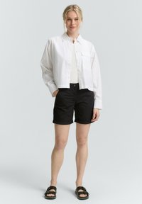 Chemise blanche cropped à boutons avec une poche poitrine unique, associée à un short noir retroussé et des sandales sombres. Design simple, coupe décontractée.