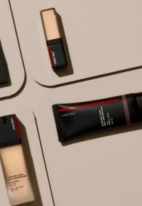 Productos de maquillaje de Shiseido sobre un fondo beige claro: dos bases líquidas en botellas de vidrio, un corrector en formato compacto y una fórmula en crema.