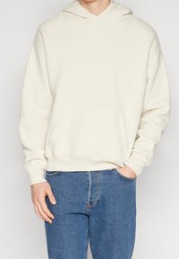 Krämfärgad hoodie i mjukt material, med ficka fram och ribbade muddärmar, i kombination med denimjeans.