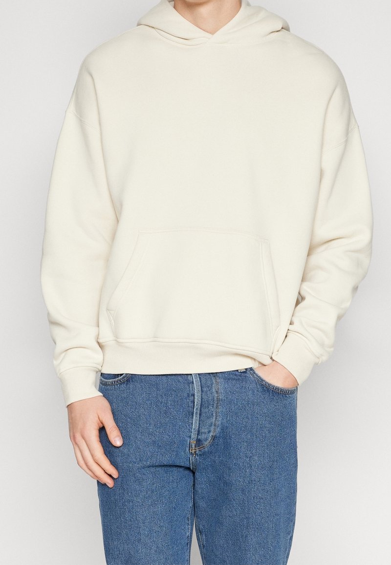 Krämfärgad hoodie i mjukt material, med ficka fram och ribbade muddärmar, i kombination med denimjeans.