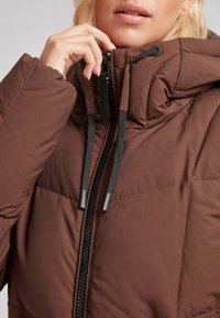 Chaqueta acolchada marrón con cuello alto y cremallera negra. El material tiene una textura suave con costuras y cordones ajustables.