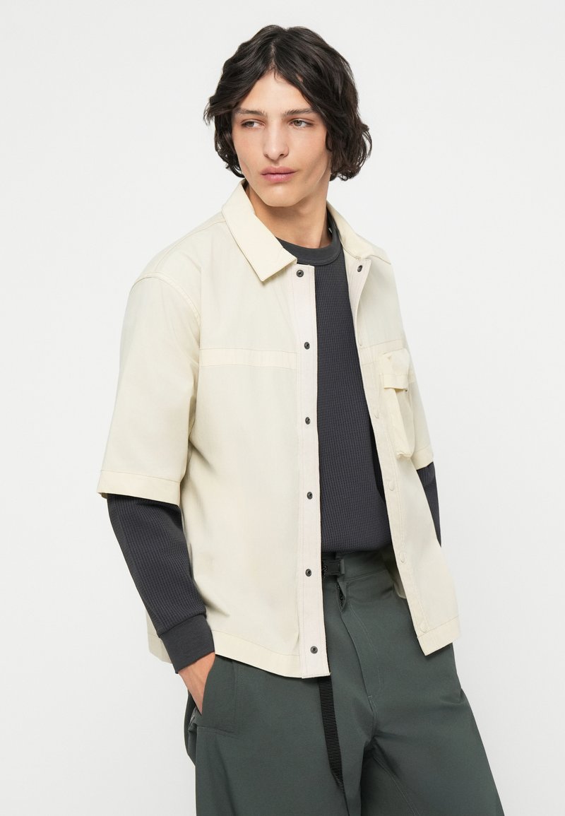 Giacca corta a maniche corte beige chiaro con colletto e bottoni neri a contrasto, indossata su una maglietta a maniche lunghe a coste scura e pantaloni verdi.