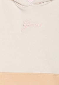 Sudadera con capucha beige con detalle en durazno claro en la parte inferior. Presenta el logo "Guess" bordado en rosa en el frente. Textura de tela suave.