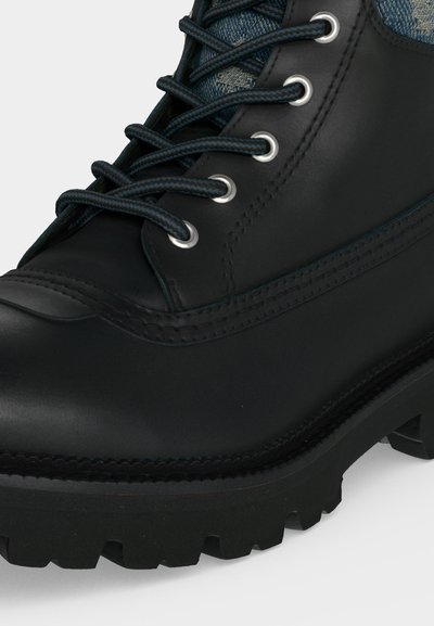 Botte en cuir noir avec semelle épaisse à crampons, œillets métalliques, lacets noirs à motifs et languette en tissu bleu visible.