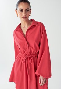 Raudona mygtukų suknelė su ilgomis, plačiomis rankovėmis ir susiaurinta juosmeniu. Pagaminta iš minkštos medžiagos su lygiu paviršiumi, su raišteliu.