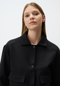 Veste noire avec un col structuré, comportant deux poches avant, des boutons mats et une texture lisse. Design épuré et minimaliste.