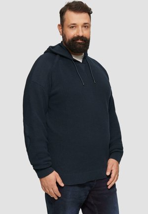 Marineblauwe gebreide hoodie met een gemêleerd patroon, geribde manchetten en een trekkoord bij de hals, gedragen met donkere jeans.