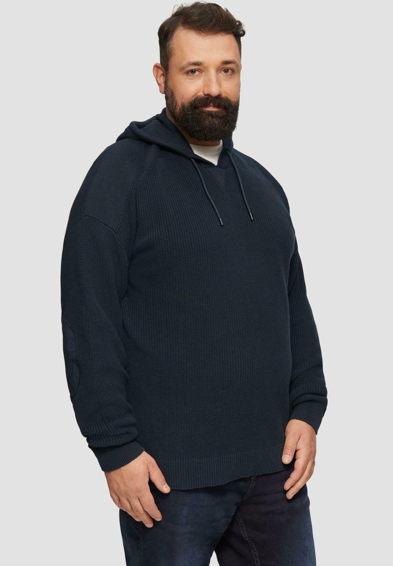 Dunkelblaue gestrickte Kapuzenjacke mit einem strukturierten Muster, gerippten Bündchen und einem Kordelzug am Hals, getragen mit dunklen Jeans.