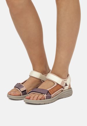 Füße in beige- und mauvefarbenen Sandalen mit offenem Zehenbereich, verstellbaren Riemen und gepolsterten Sohlen vor einfachem hellem Hintergrund.