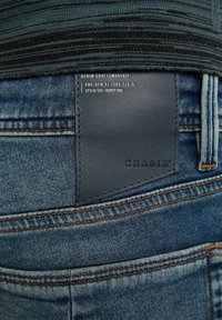 Zadní pohled na modré džínové kalhoty s hnědým prošíváním a černou koženou nášivkou s nápisem "CHASIN" a "DENIM CRAFTSMANSHIP."