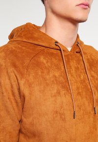 Hoodie de ante marrón con un acabado texturizado, que presenta una capucha con cordones y ojales de tono plateado. La tela parece suave y ligeramente usada.