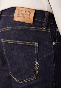 Tumed denim jeans, mille pruuni nahklogo plaastriga vöökohtades, kontrastsete kollaste õmblustega ja ainulaadse õmmeldud detailiga tagakotil.