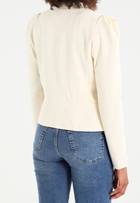 Blazer aderente color crema con spalle strutturate, tessuto testurizzato e vita a peplo, abbinato a jeans in denim blu.