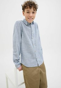 Camicia a righe blu e bianche con maniche lunghe, colletto e bottoni marroni; indossata con pantaloni corti beige su uno sfondo neutro.