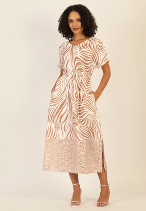 Frau mit lockigem Haar, die ein midi-kleid mit beigen und weißen Mustern, kurzen Ärmeln und seitlichen Schlitzen trägt, steht mit den Händen in den Taschen.