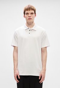 Bílá bavlněná polo košile s krátkým rukávem, klasickým límečkem a třemi černými knoflíky. Hladká textilie, pohodlný střih a minimalistický design.