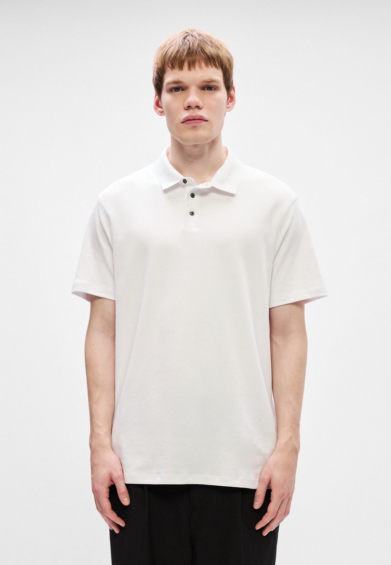 Polo en coton blanc à manches courtes, col classique et trois boutons noirs. Texture lisse, coupe décontractée et design minimal.