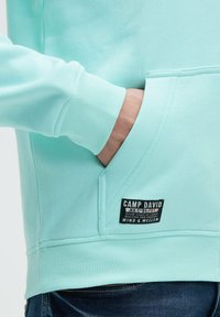 Felpa verde menta con polsini e orlo a costine, tasca frontale e etichetta con logo e testo. Tessuto morbido e design casual.