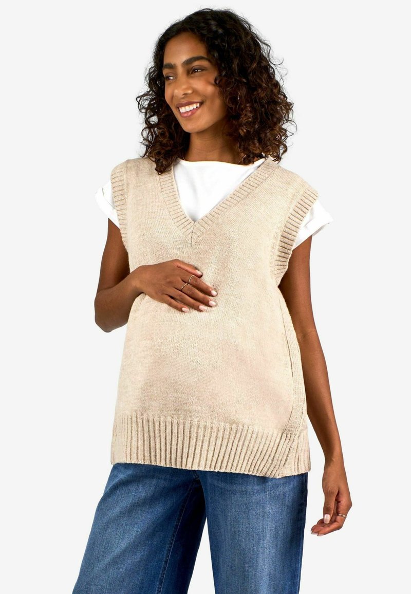Beige kudumisest ilma varrukateta sviiter V-kaelusega, ribidega ääristatud ja lõdva lõikega, koos siniste denim teksadega.