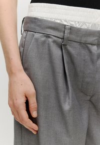 Graue Hosen mit einem gefalteten Design, strukturiertem Stoff und einem kontrastierenden weißen elastischen Bund. Die Hand ruht auf dem Bund für besseren Halt.