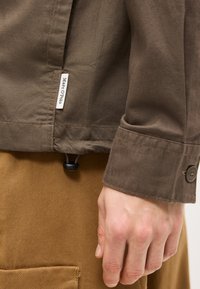 Καφέ μανσέτα σακακιού με κουμπί, ετικέτα Marc O’Polo κοντά στο τελείωμα και μπεζ παντελόνι με ορατή λεπτομέρεια τσέπης στο χέρι ενός ατόμου.