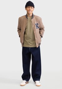 Brown houndstooth bomber jacket with navy accents, olive green T-shirt, dark blue wide-leg jeans, and white sneakers. Black knit hat.