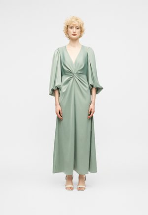 Kvinne med kort, krøllet blondt hår iført en lys grønn lang kjole med puffede ermer og beige åpen tå-hæler, stående foran en ensfarget hvit bakgrunn.