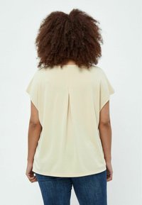 Beige Oberteil mit kurzen Ärmeln und lässiger Passform, mit Faltendetails im Rücken und weicher Textur. Kombiniert mit blauen Jeans.