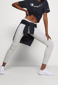 Camiseta corta de color azul oscuro con nudo en la parte delantera, leggings grises y zapatillas deportivas blancas. El atuendo incluye un sudadera color marino atada a la cintura.
