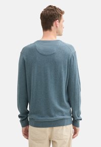 Maglione blu a maglia con maniche lunghe e collo rotondo, caratterizzato da una texture liscia e polsini e orlo a coste. Cuciture visibili sulle spalle.