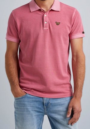 Mężczyzna w różowej koszulce polo z krótkim rękawem, ozdobionej małym haftowanym logo ptaka, oraz w jasnoniebieskich dżinsach, z jedną ręką w kieszeni.