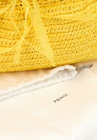 Sac à main tissé jaune posé sur une housse anti-poussière blanche de la marque Fendi avec un cordon gris sur une surface blanche.