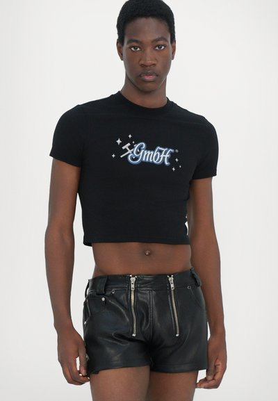 Tricou scurt negru cu grafică „GmbH” în albastru și alb, asortat cu şorturi din piele neagră cu fermoare metalice și o ajustare strânsă.