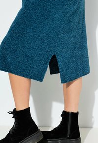 Gonna lavorata a maglia color turchese con una spaccatura laterale, abbinata a stivaletti neri con lacci e suola testurizzata, dotati di dettaglio con zip.