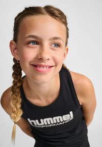 Svart ärmlös linne med vit "hummel"-logga; flätat hår; ljus hy; leende uttryck; mjuk, slät textur.