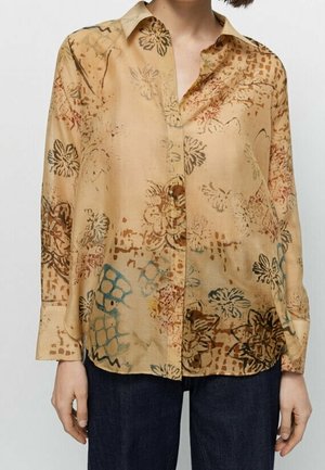 Beige button-up blouse with abstract floral and geometric patterns in brown, blue, and red, worn with dark blue pants.