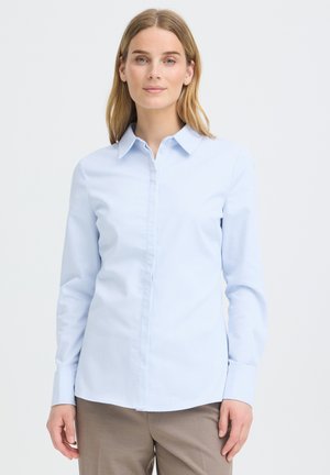 Femme aux cheveux châtain clair portant une chemise bleue claire boutonnée et un pantalon beige, debout devant un fond blanc uni.