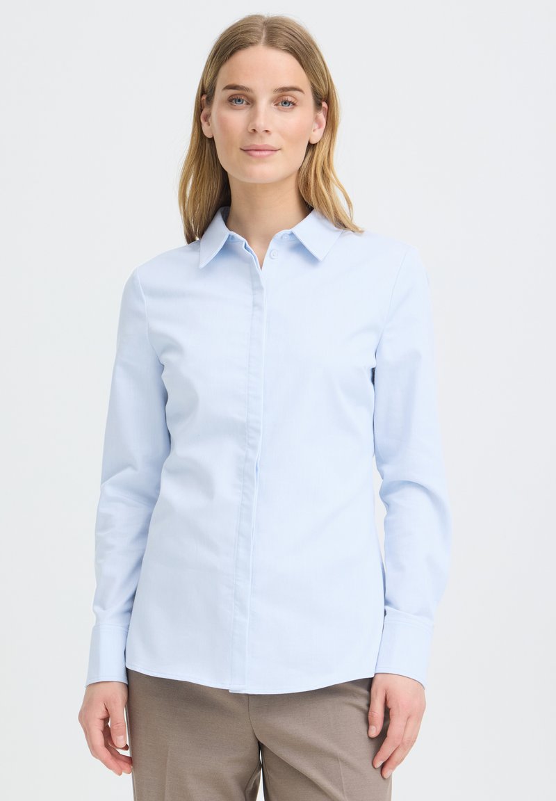 Femme aux cheveux châtain clair portant une chemise bleue claire boutonnée et un pantalon beige, debout devant un fond blanc uni.