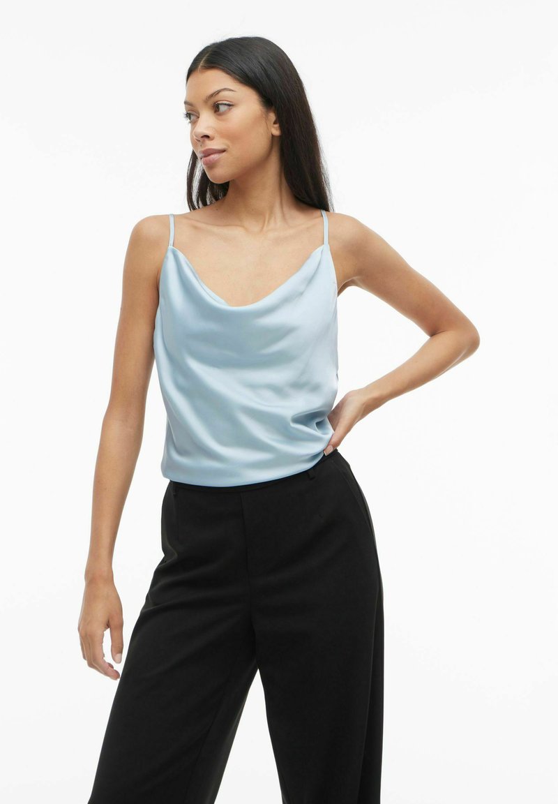 Svijetloplava satenasta bluzica s naglašenim izrezom i tankim naramenicama, u kombinaciji s crnim širokim hlačama koje imaju glatku teksturu i opušteni kroj.