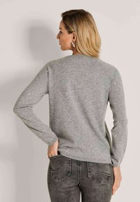 Frau in einem grauen Pullover mit kleinen Verzierungen an den Ärmeln und ausgewaschenen schwarzen Jeans, die mit dem Rücken zur Kamera steht.