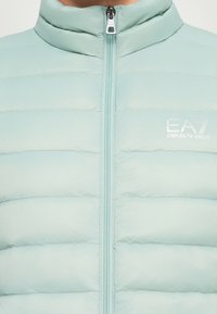 Giacca imbottita azzurro chiaro con zip frontale e logo Emporio Armani EA7 sul petto, caratterizzata da dettagli di cuciture imbottite orizzontali.