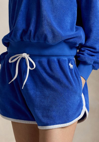 Άτομο που φορά μπλε σορτς από περκάλι με λευκό κορδόνι και αντίστοιχο μπλε φούτερ, με το χέρι στην τσέπη, στέκεται μπροστά σε ουδέτερο φόντο.