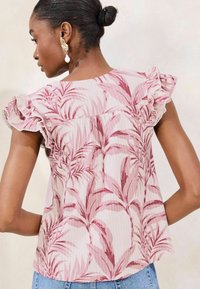 Light pink short-sleeve top with ruffled accents, featuring a palm leaf pattern in deeper pink. Textured fabric with a relaxed fit.