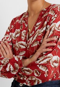 Rode bloemenblouse met een V-hals. Bevat witte en bruine bloemmotieven. Lange mouwen met knoopmanchetten en een zachte textuur.