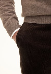 Maglione marrone a maglia con polsini a coste, abbinato a pantaloni in corduroy marrone scuro con righe verticali. Mano appoggiata in tasca.